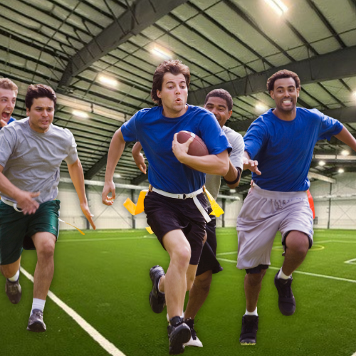 Football players sprinting on an indoor turf field, with a blue-shirted runner carrying the ball.