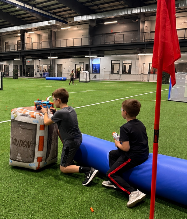 Two boys at an indoor sports field, one kneeling behind a blue barrier and a red flag in the foreground.