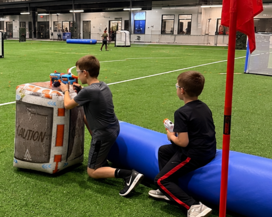 Two children kneel by a foam obstacle on an indoor turf field, one aiming a toy blaster.