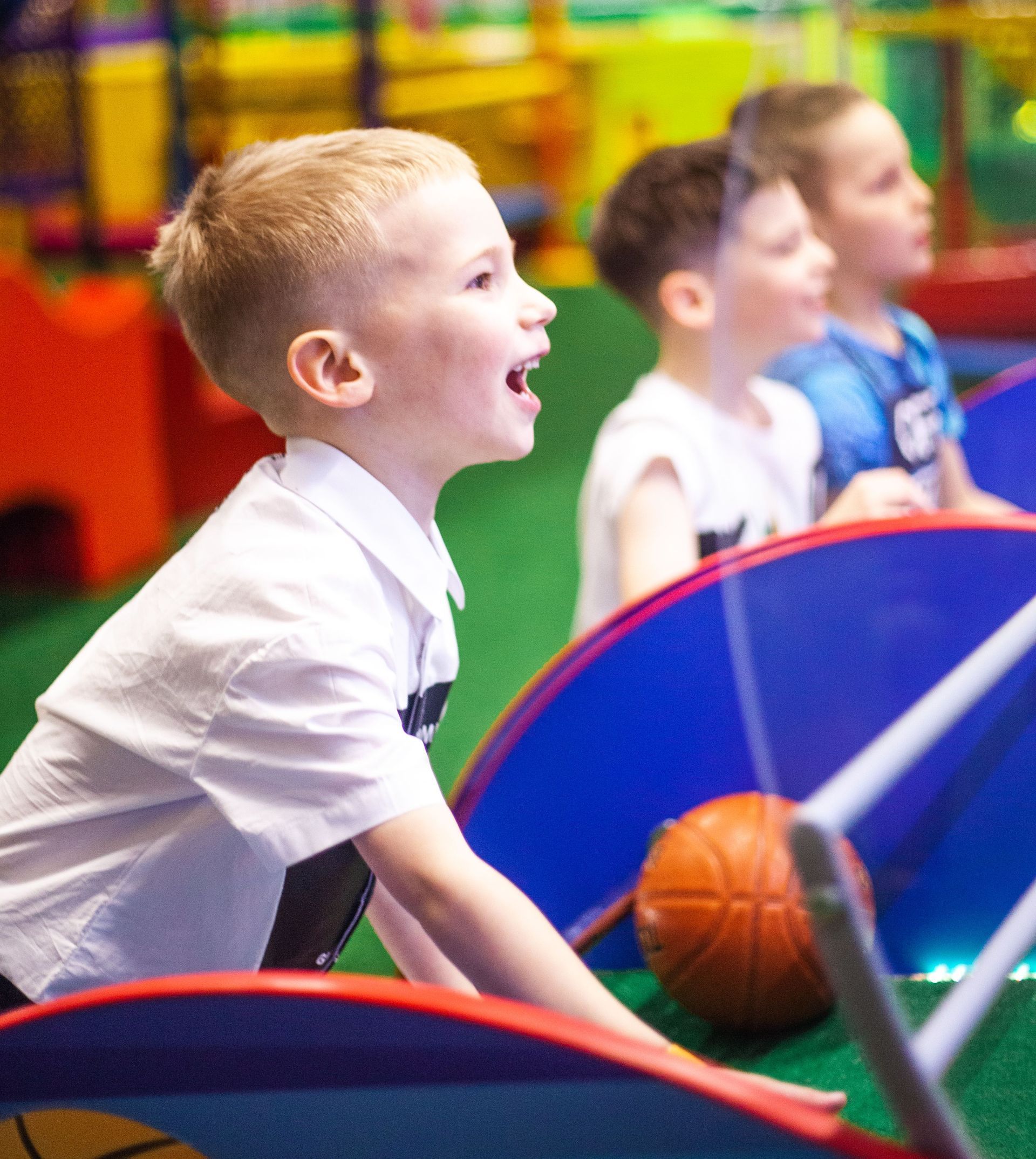 Children playing in an indoor arcade, leaning over colorful game panels with a basketball and bright play equipment.