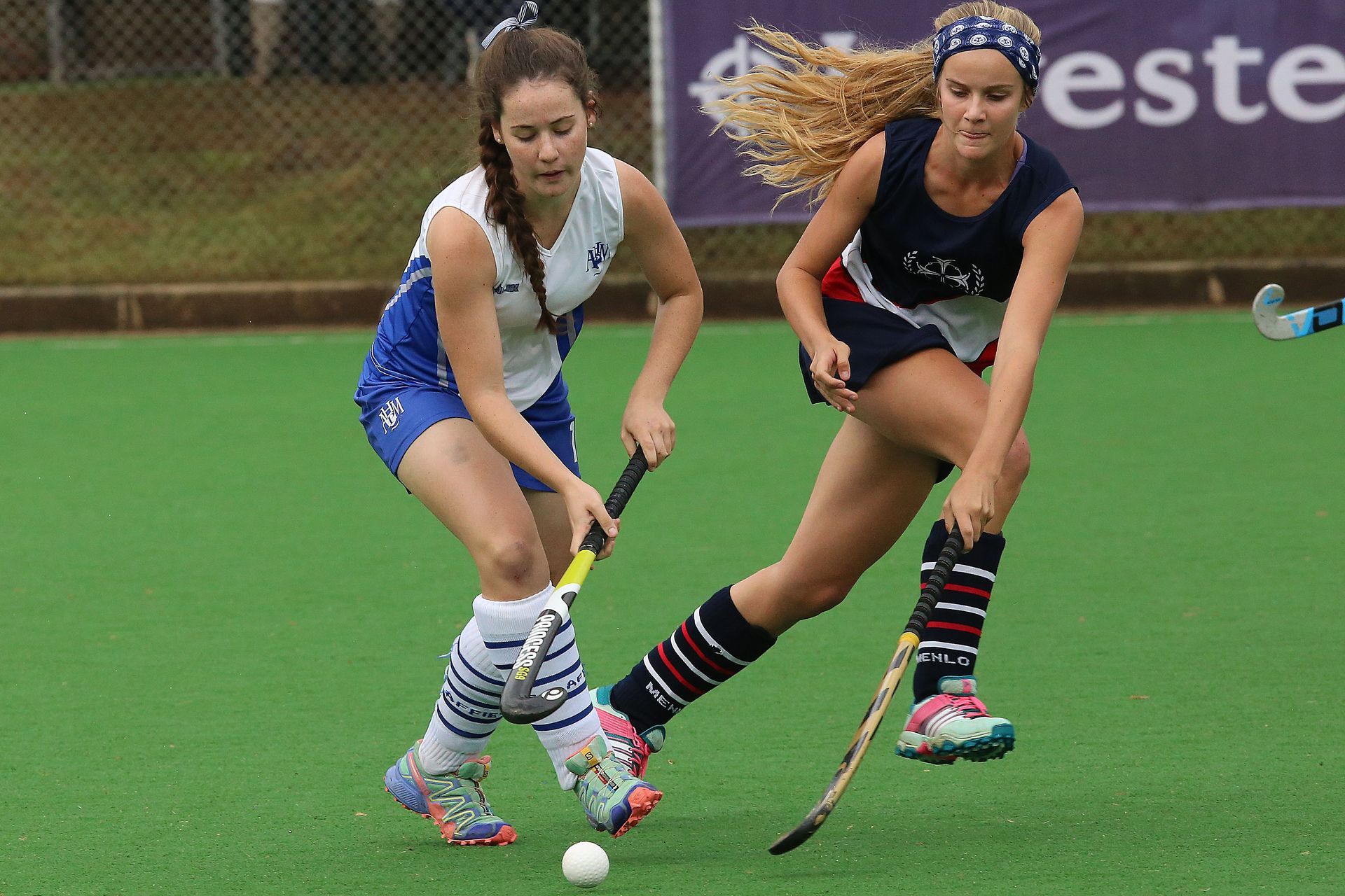 Two field hockey players in a match, chasing the ball on a green turf field