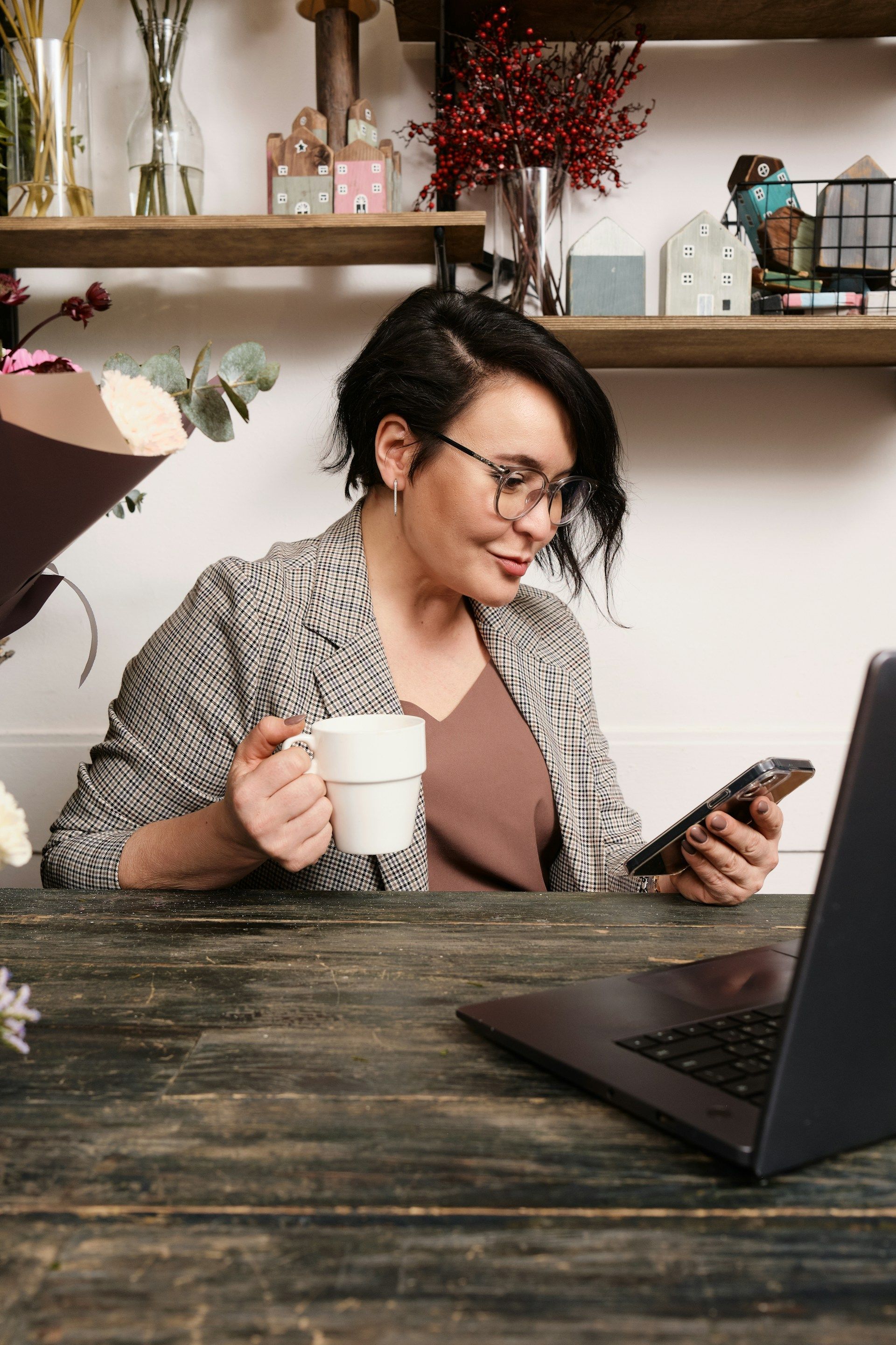 A woman is sitting at a table holding a cup of coffee and looking at her phone.