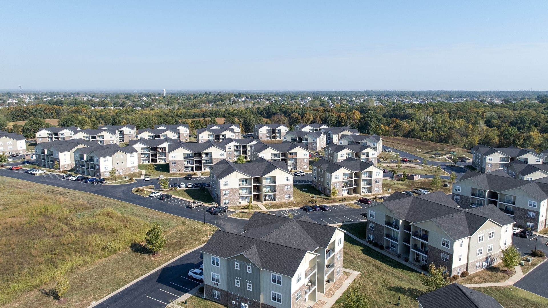 Aerial view of a suburban apartment complex with gray buildings, parking lots, and surrounding open land.