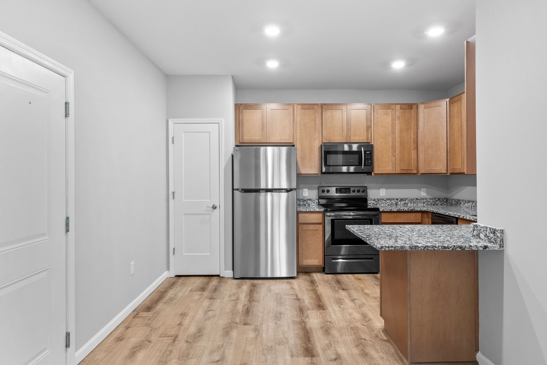 Empty modern kitchen with stainless appliances, wood cabinets, and granite countertops on light wood flooring