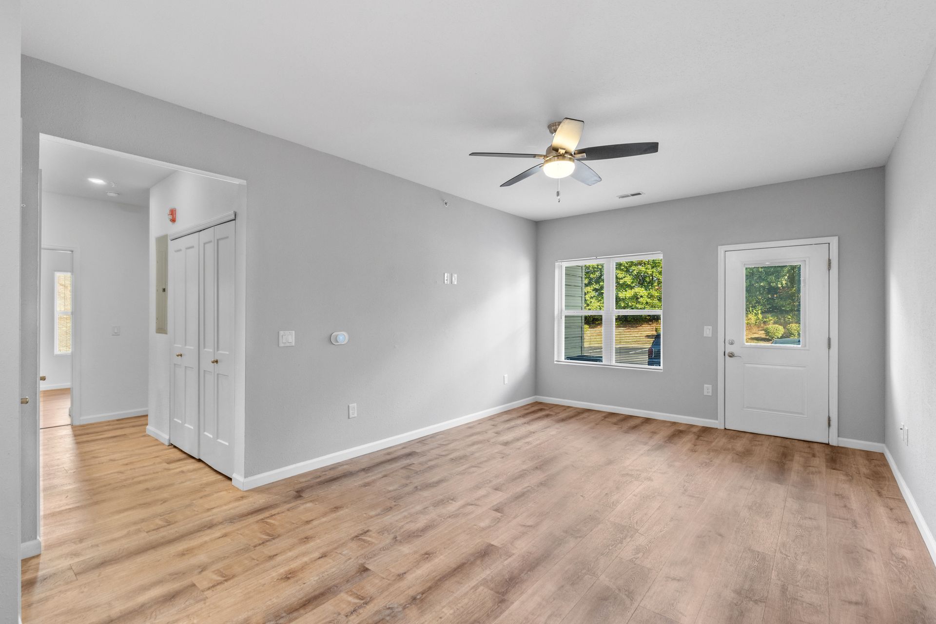 Empty gray living room with wood floors, ceiling fan, windows, and white front door