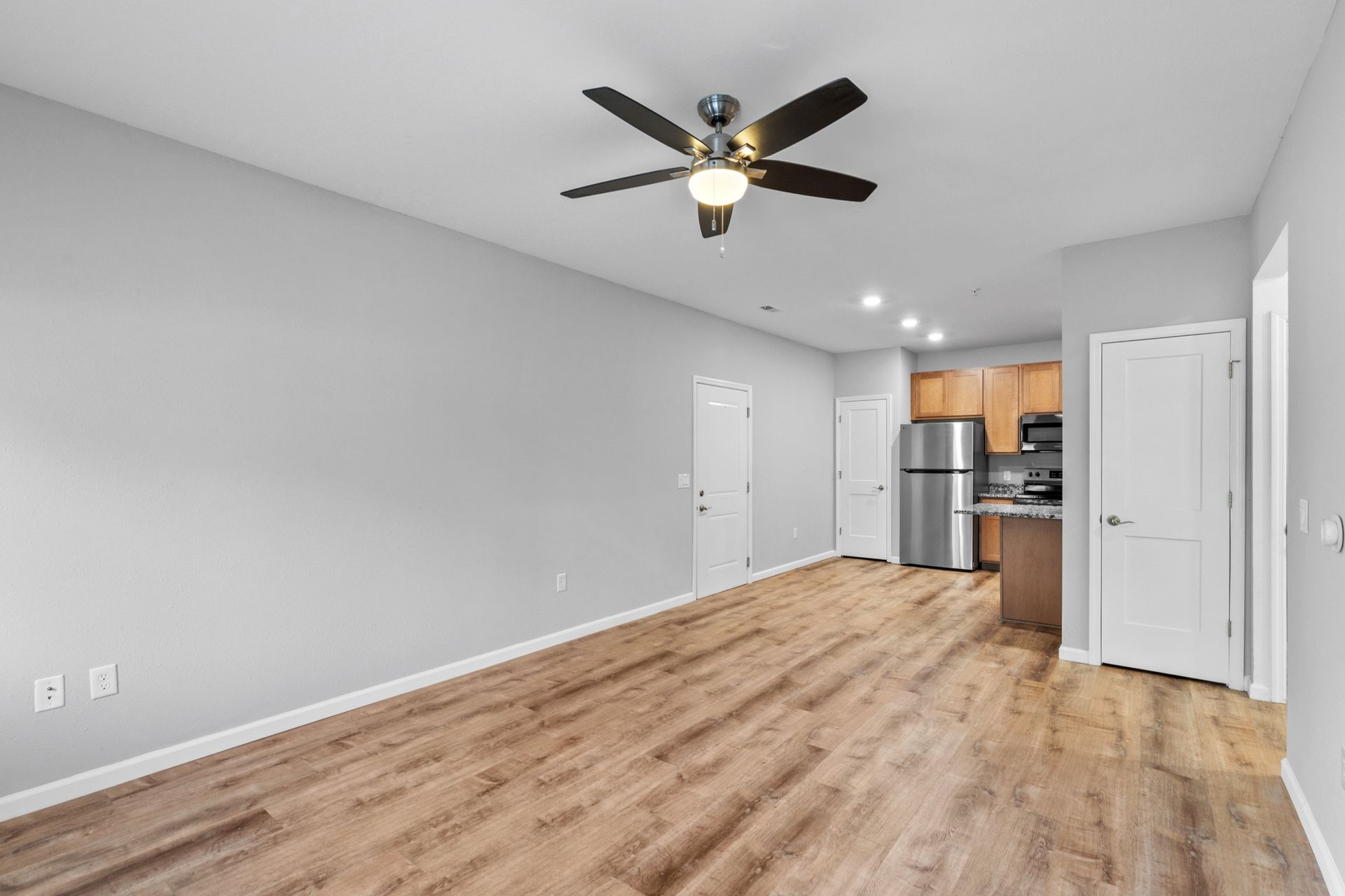 Empty modern living room with wood floors, ceiling fan, and a small kitchen area in the back