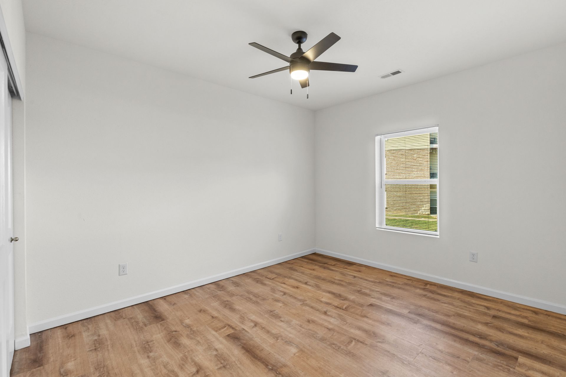 Empty white bedroom with wood-look floor, ceiling fan, and a window with blinds.