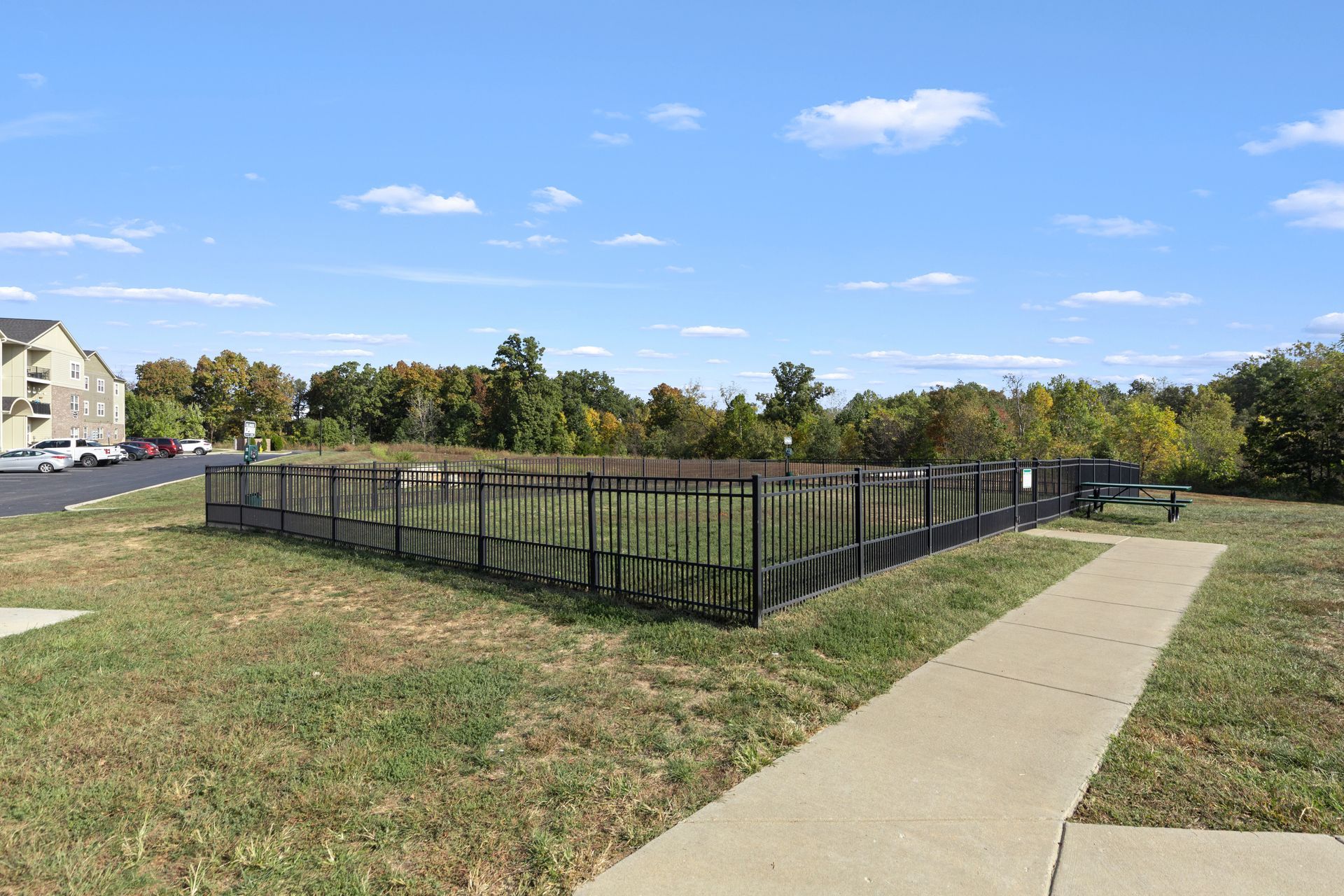 Fenced grass enclosure beside a sidewalk in a suburban park, with trees and apartments in the background