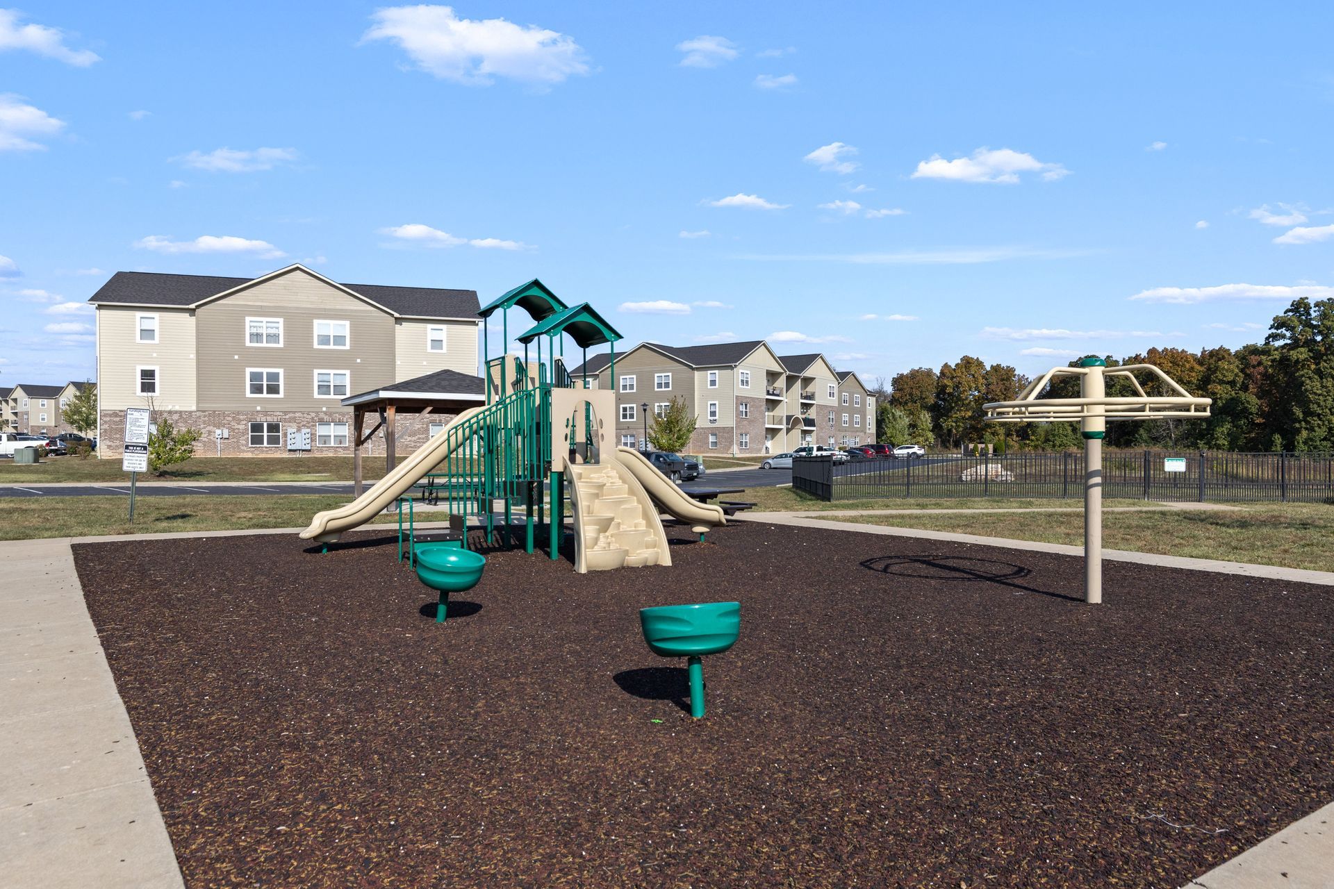 Playground with green play structure and tan slides on dark mulch, surrounded by apartment buildings under a blue sky