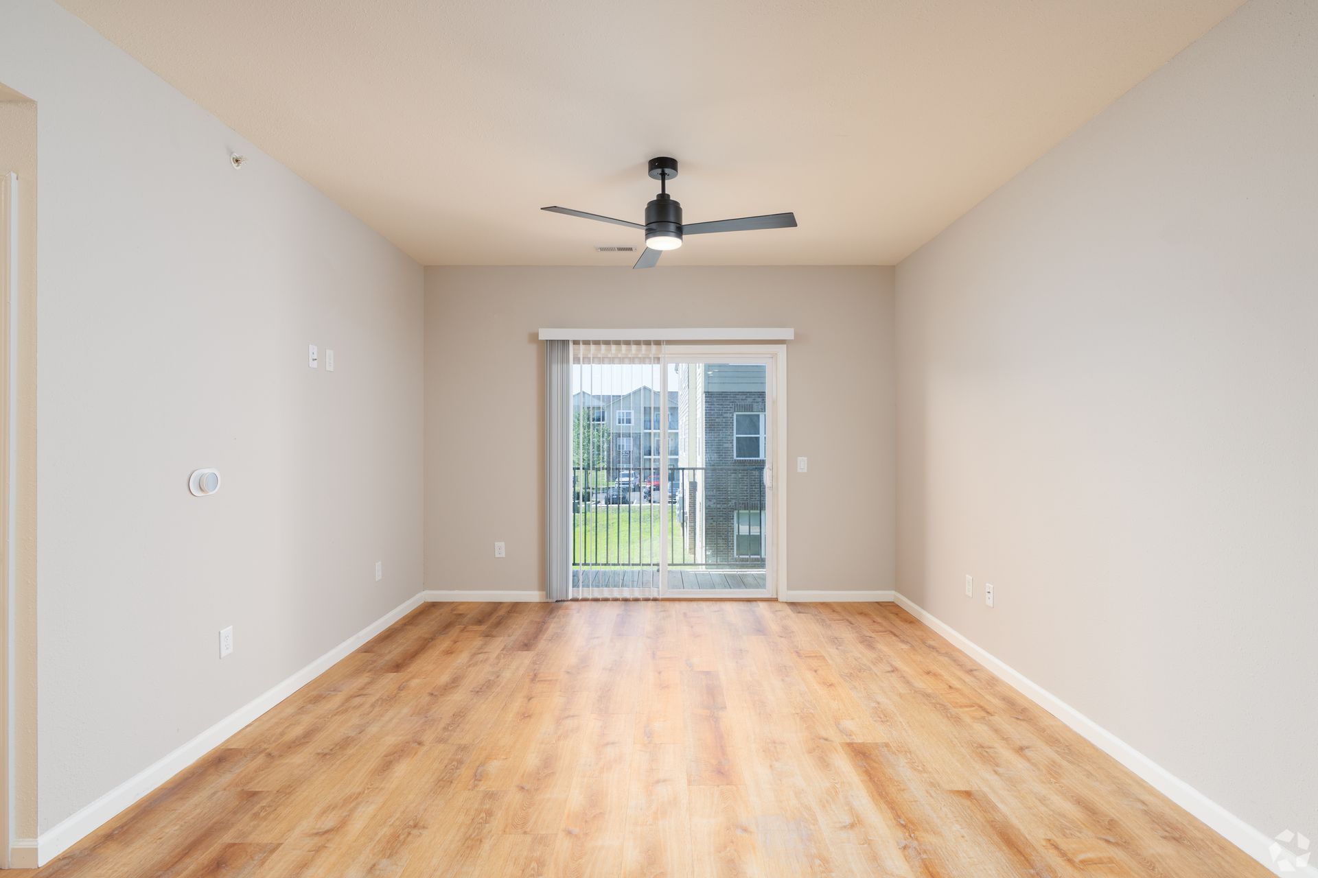 Empty living room with light wood floors, ceiling fan, and sliding glass doors to a balcony.