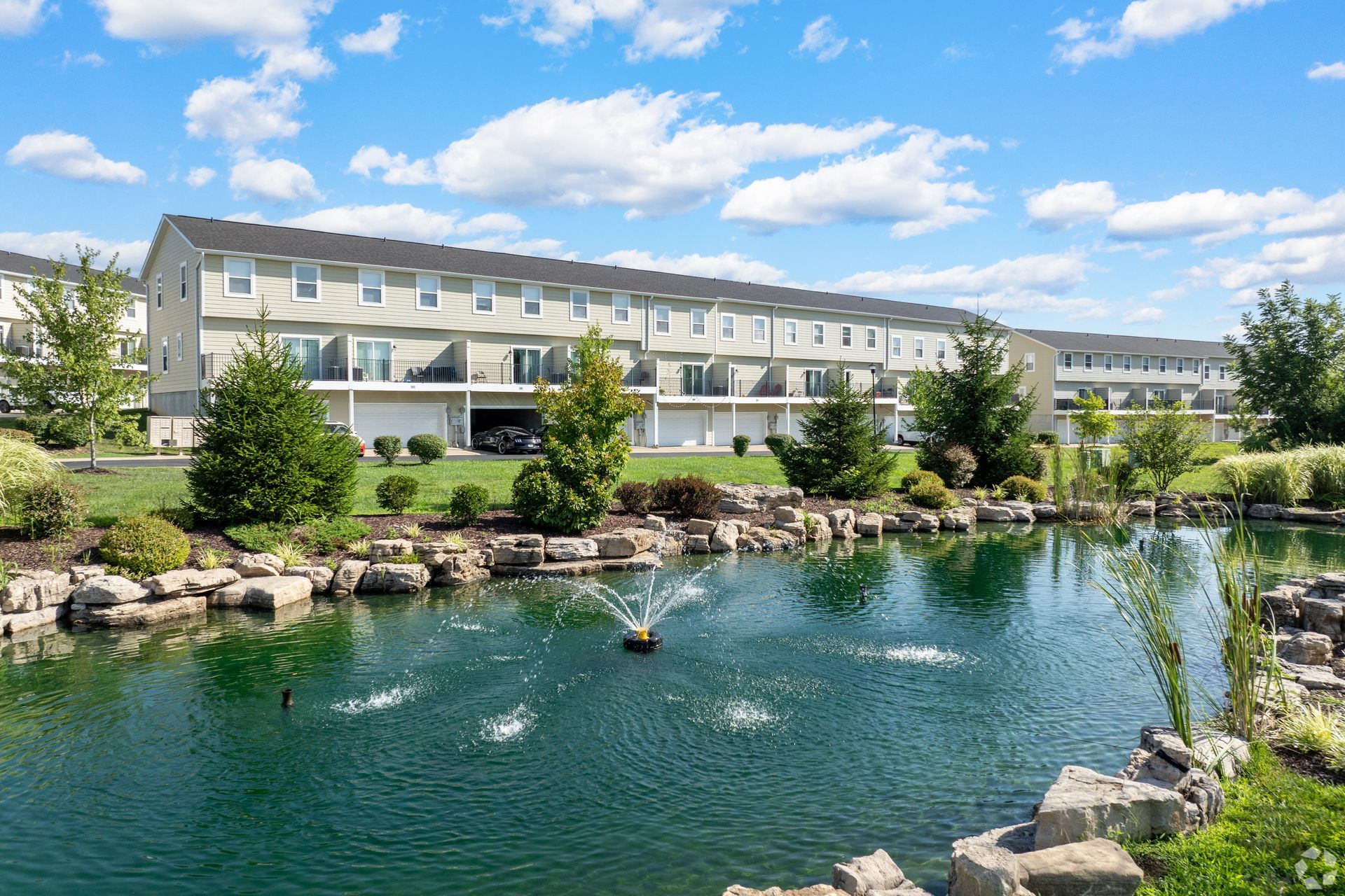 Lakeside resort buildings under a blue sky, with a pond, rocks, and ducks in the foreground