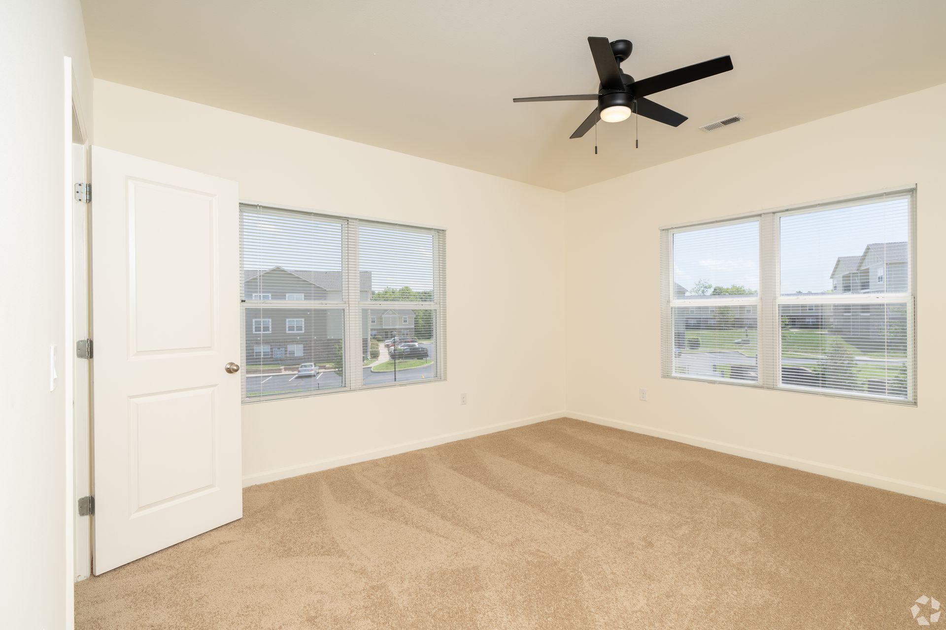 Empty beige bedroom with carpet, white walls, two windows, and a black ceiling fan