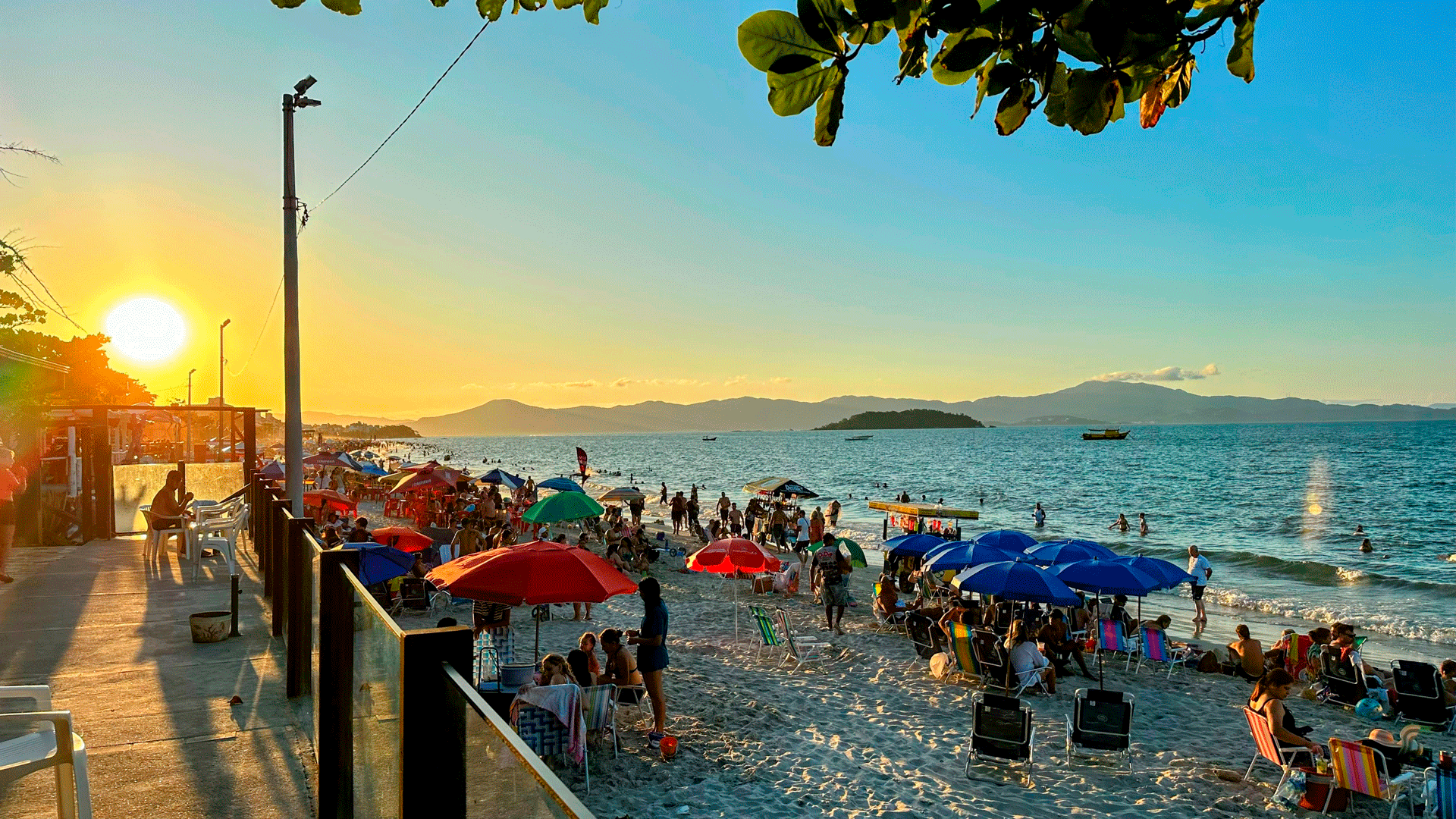 Cena de praia ao pôr do sol; pessoas sob guarda-sóis e ao longo da costa, água e ilha ao longe.