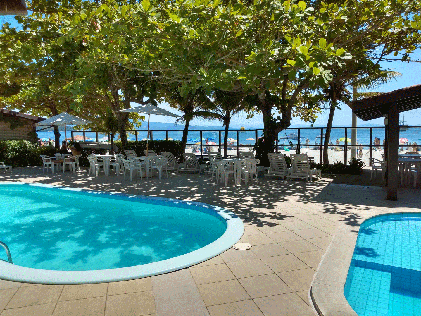 Vista da praia com piscina, mesas brancas, árvores, água azul e céu.