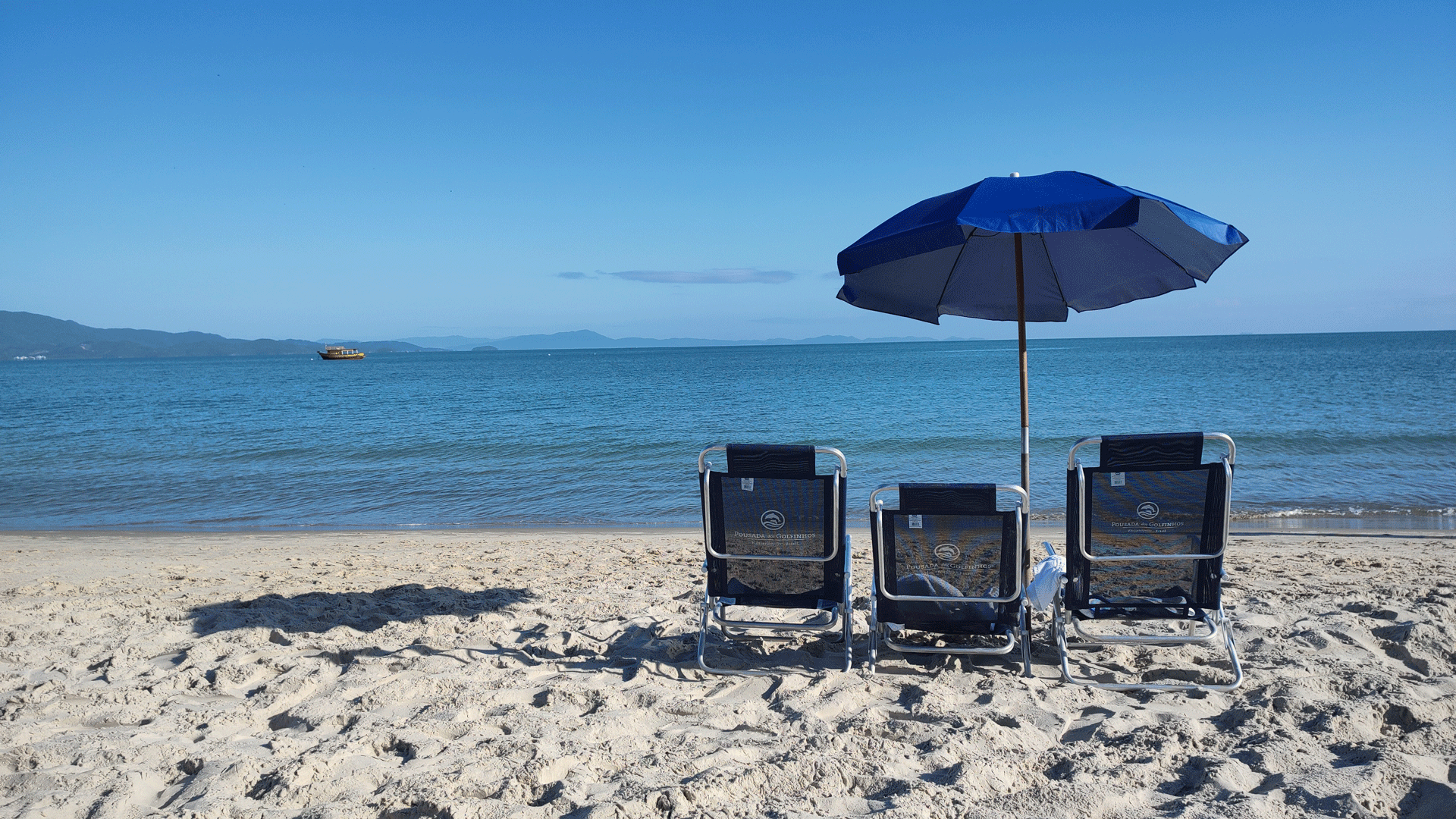 Três cadeiras de praia sob um guarda-sol azul em uma praia de areia, com o oceano ao fundo.