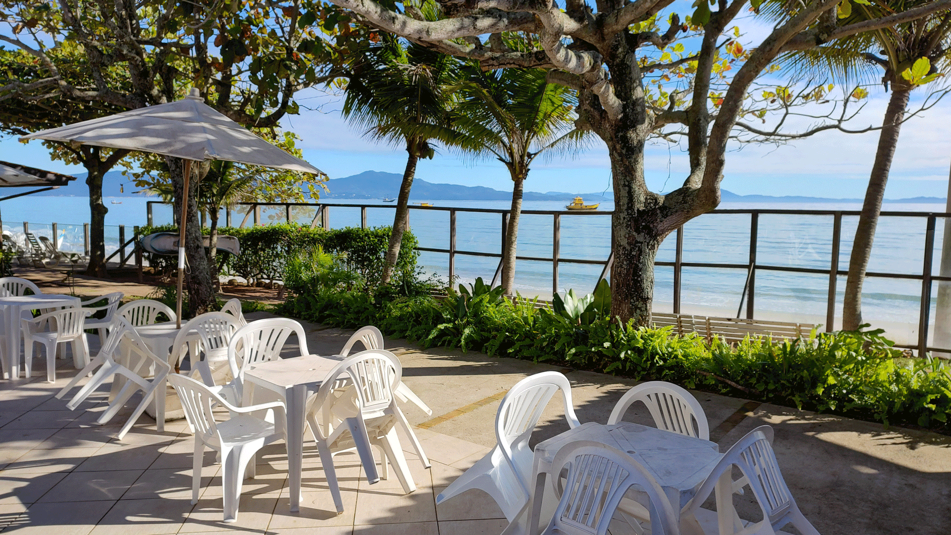 Mesas e cadeiras em um pátio com vista para a praia e o oceano, com árvores.