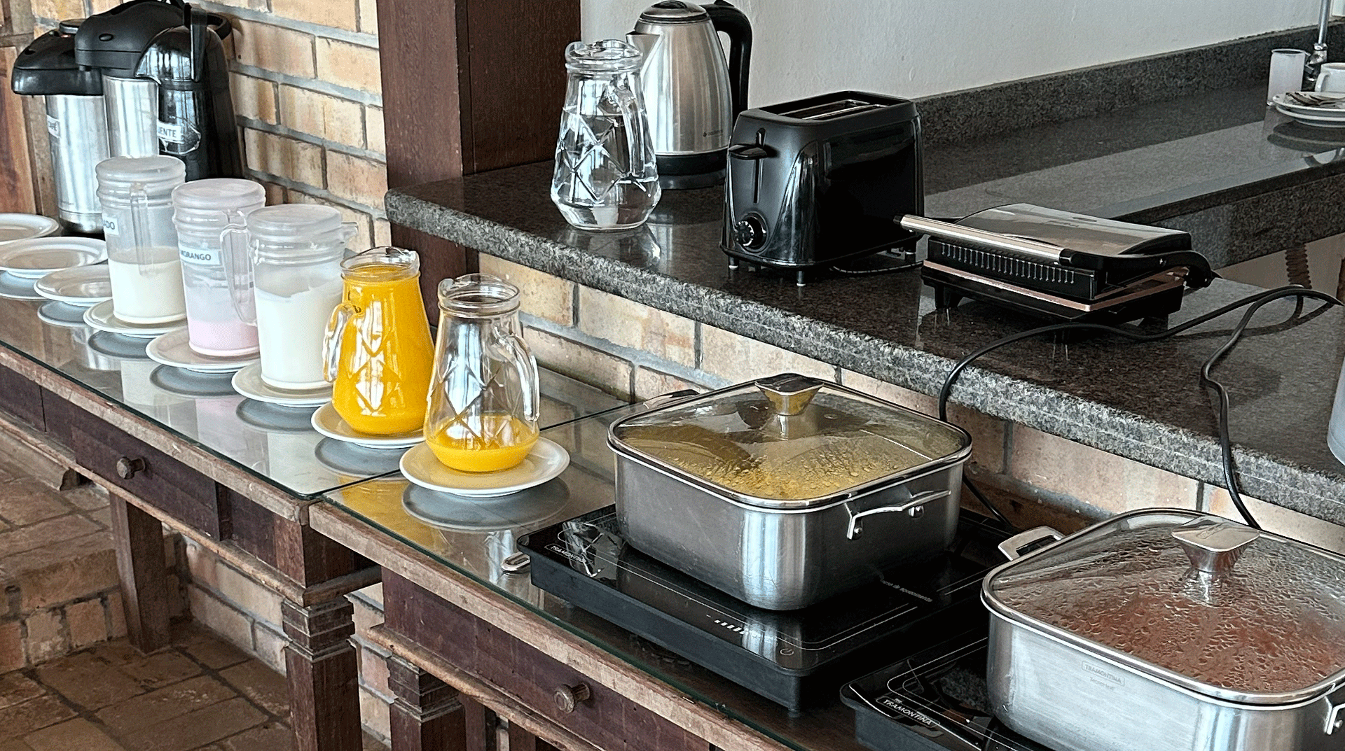 Buffet de café da manhã com bebidas, torradeira e recipientes de comida quente no balcão.