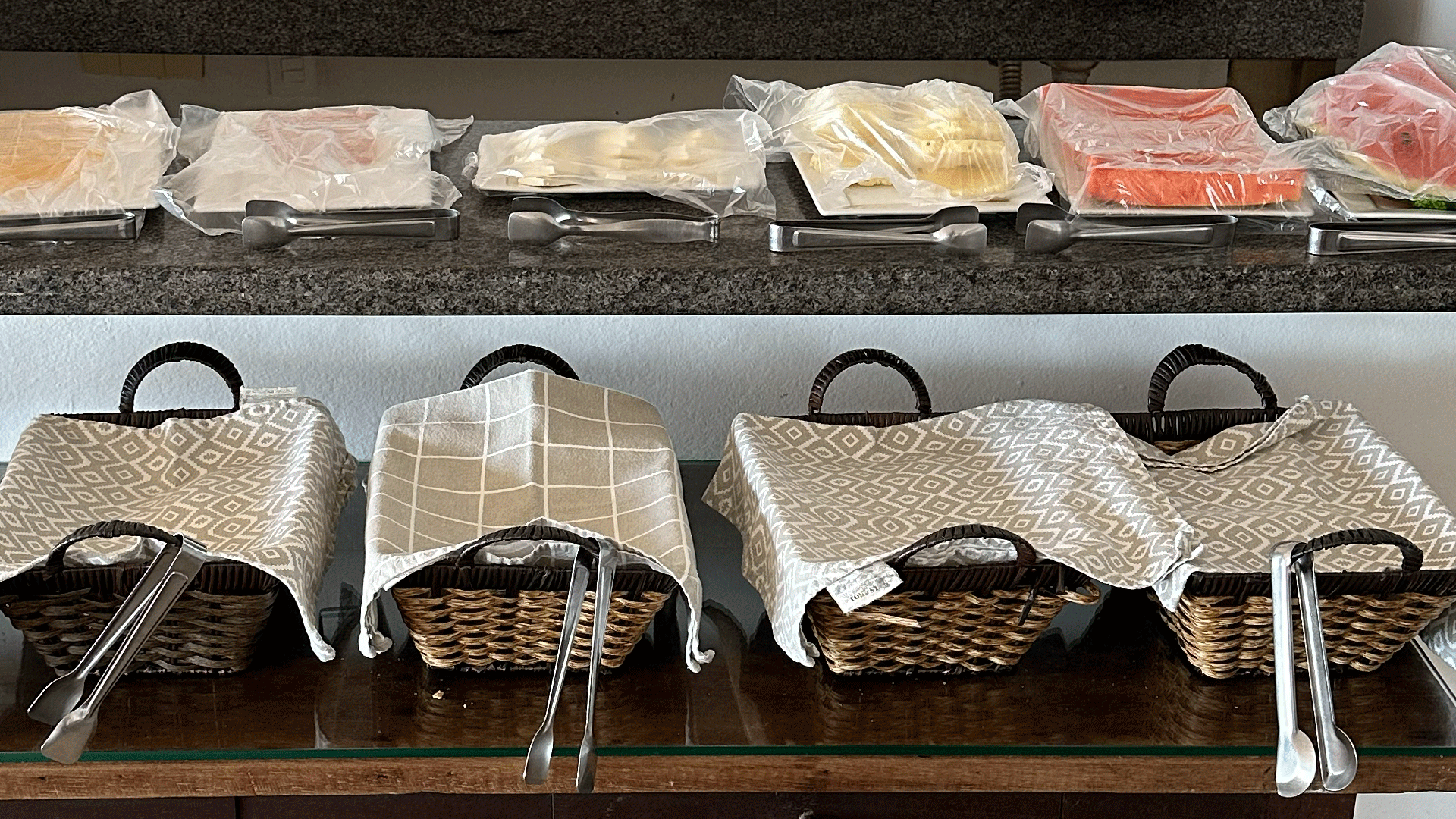 Buffet com alimentos embalados na prateleira superior, cestos de pão cobertos embaixo e pinças.