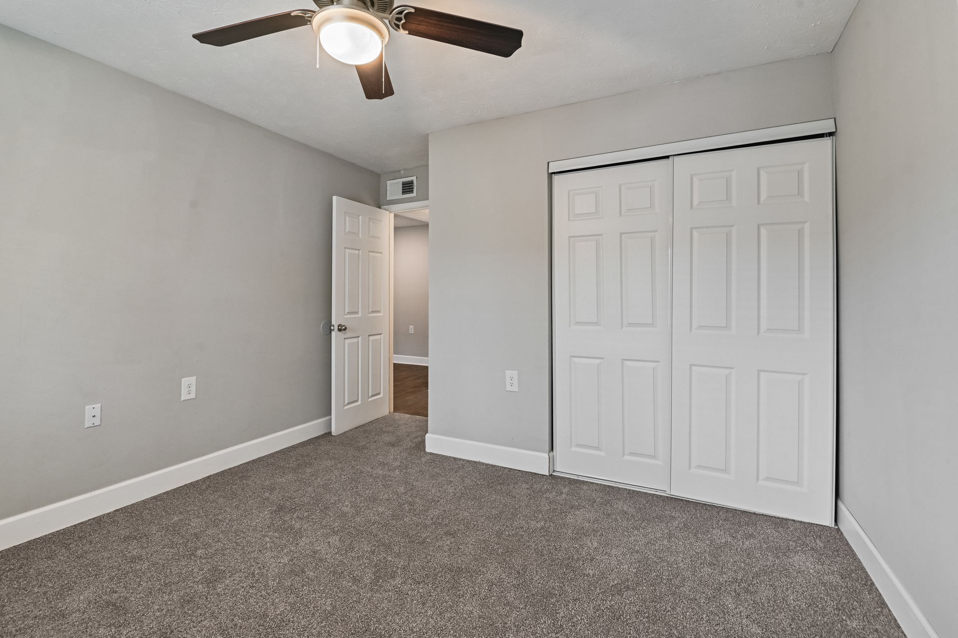 A bedroom with light gray walls, gray carpet, a white ceiling fan, an open doorway, and white paneled sliding closet doors.