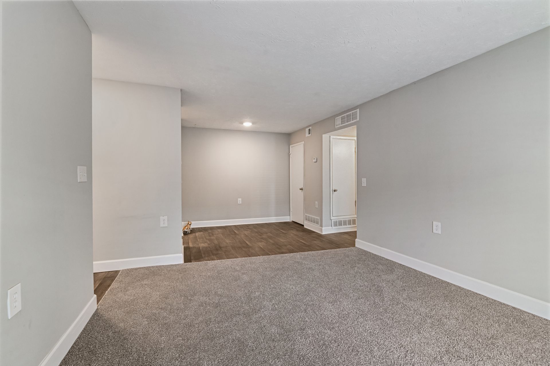 A bright, empty room with light gray walls, white trim, and light gray carpet, featuring an adjoining carpeted alcove.