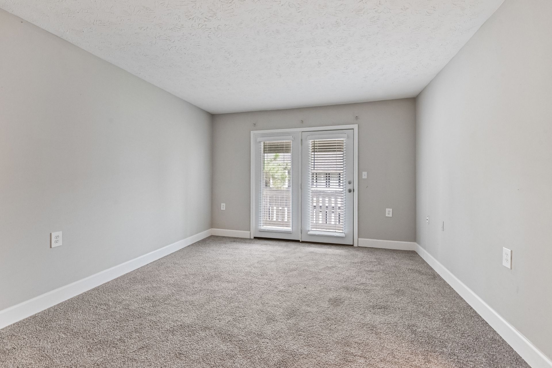 An empty room with light gray walls, gray carpeting, a white popcorn ceiling, and glass double doors leading outside.