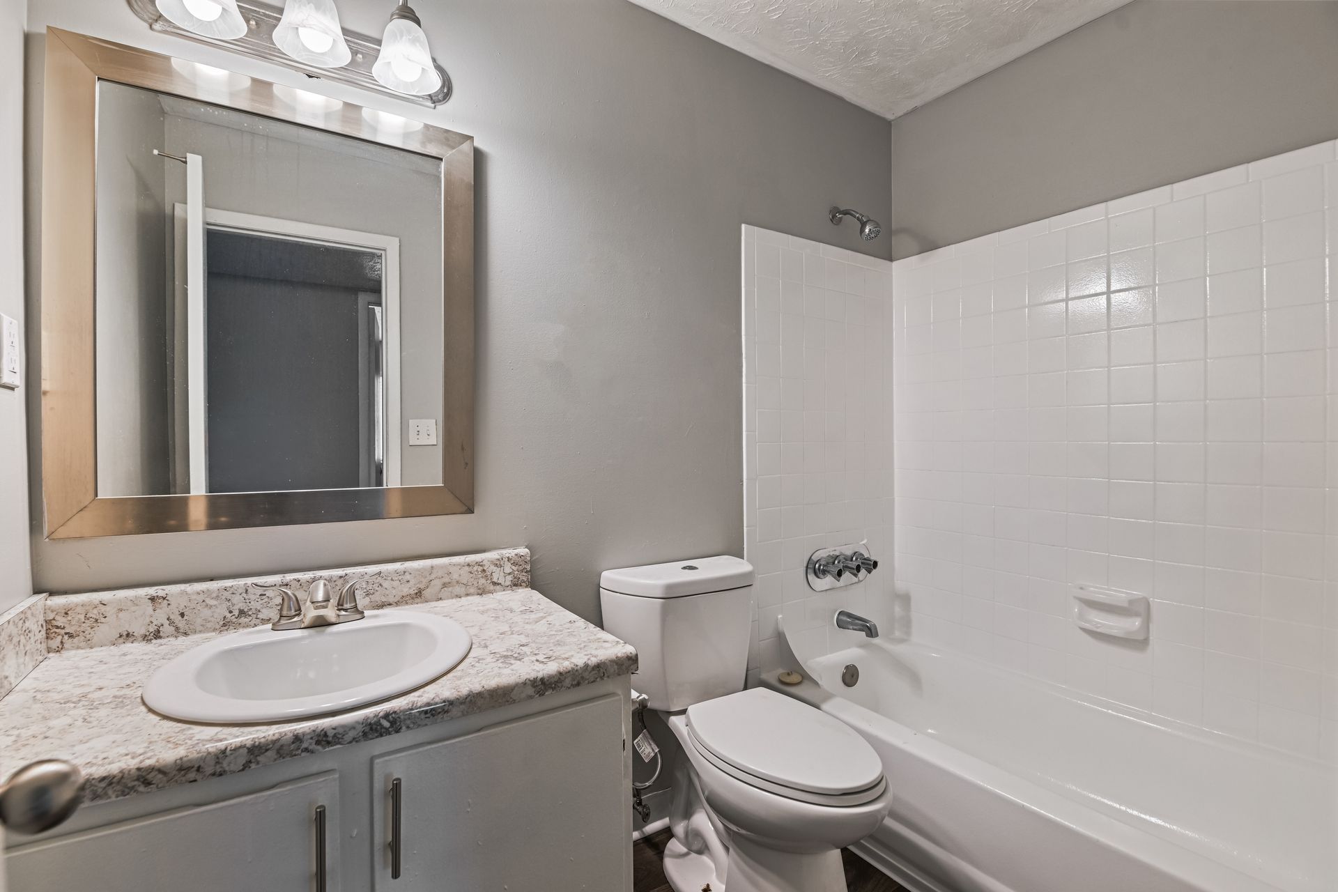 A bathroom featuring a light granite vanity, a framed mirror, a toilet, and a white tiled bathtub shower combination.