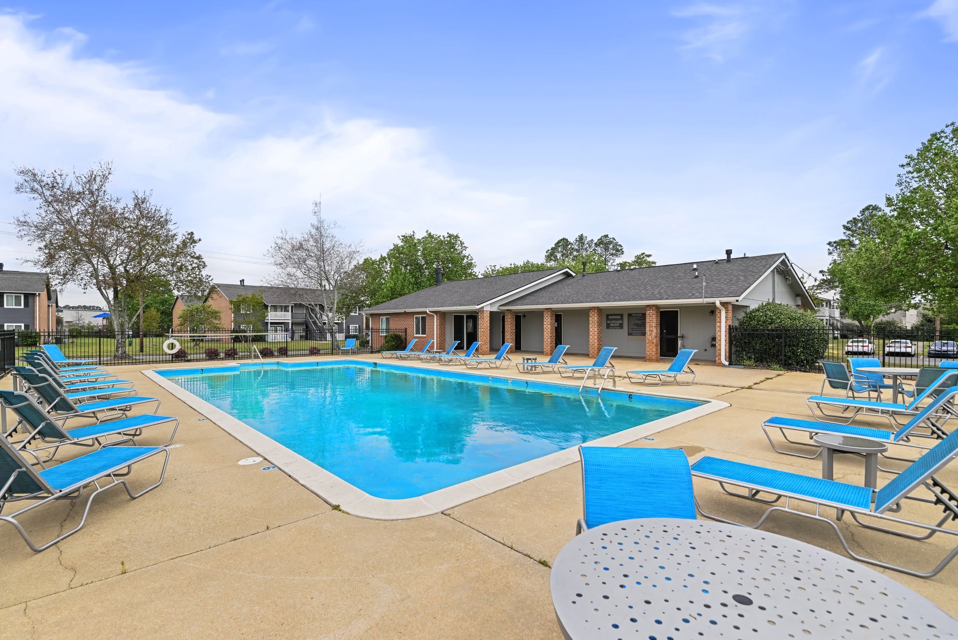 An outdoor swimming pool at an apartment complex, surrounded by lounge chairs on a deck with a clubhouse in the background.
