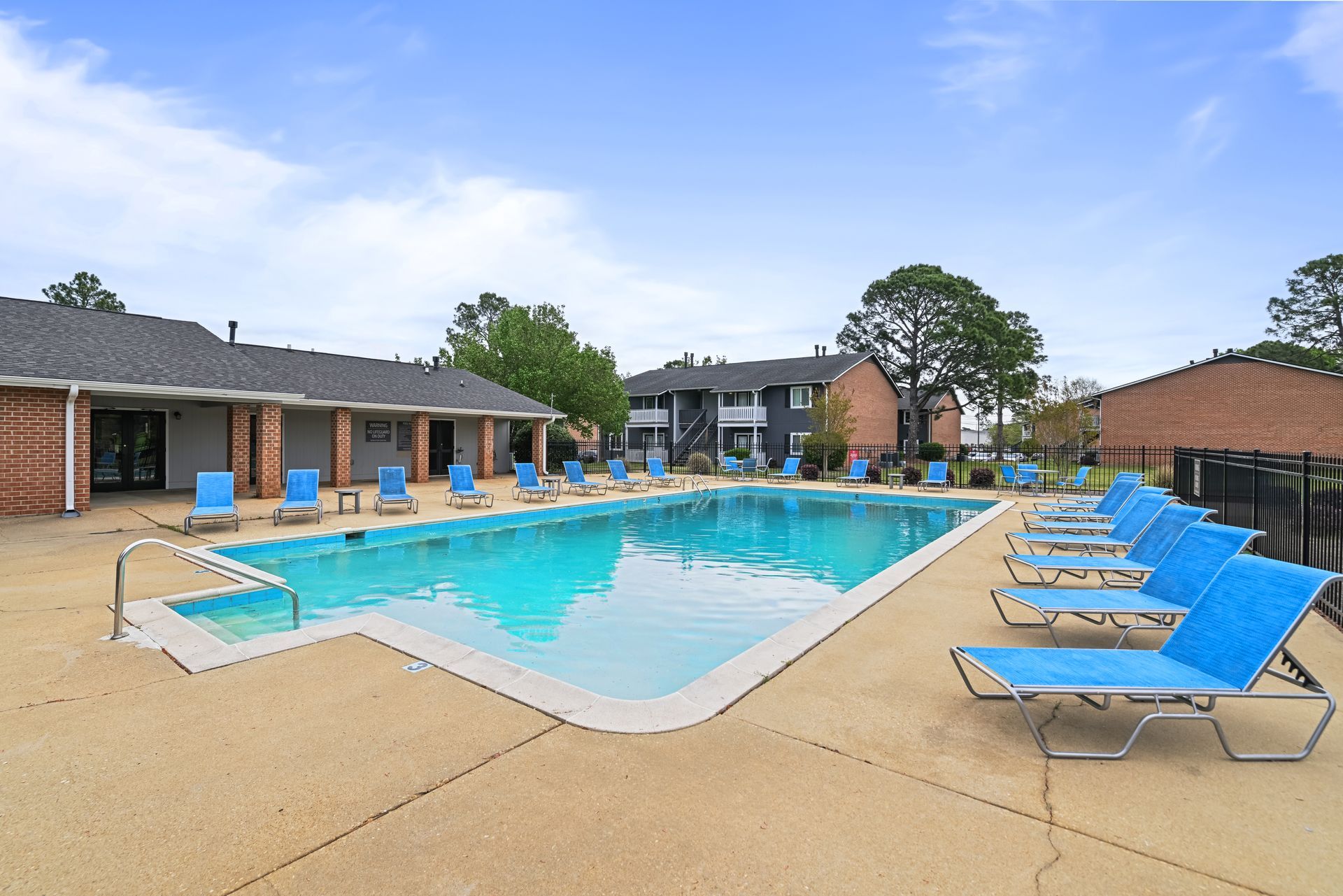 An outdoor swimming pool surrounded by a concrete patio with several blue lounge chairs under a clear blue sky.