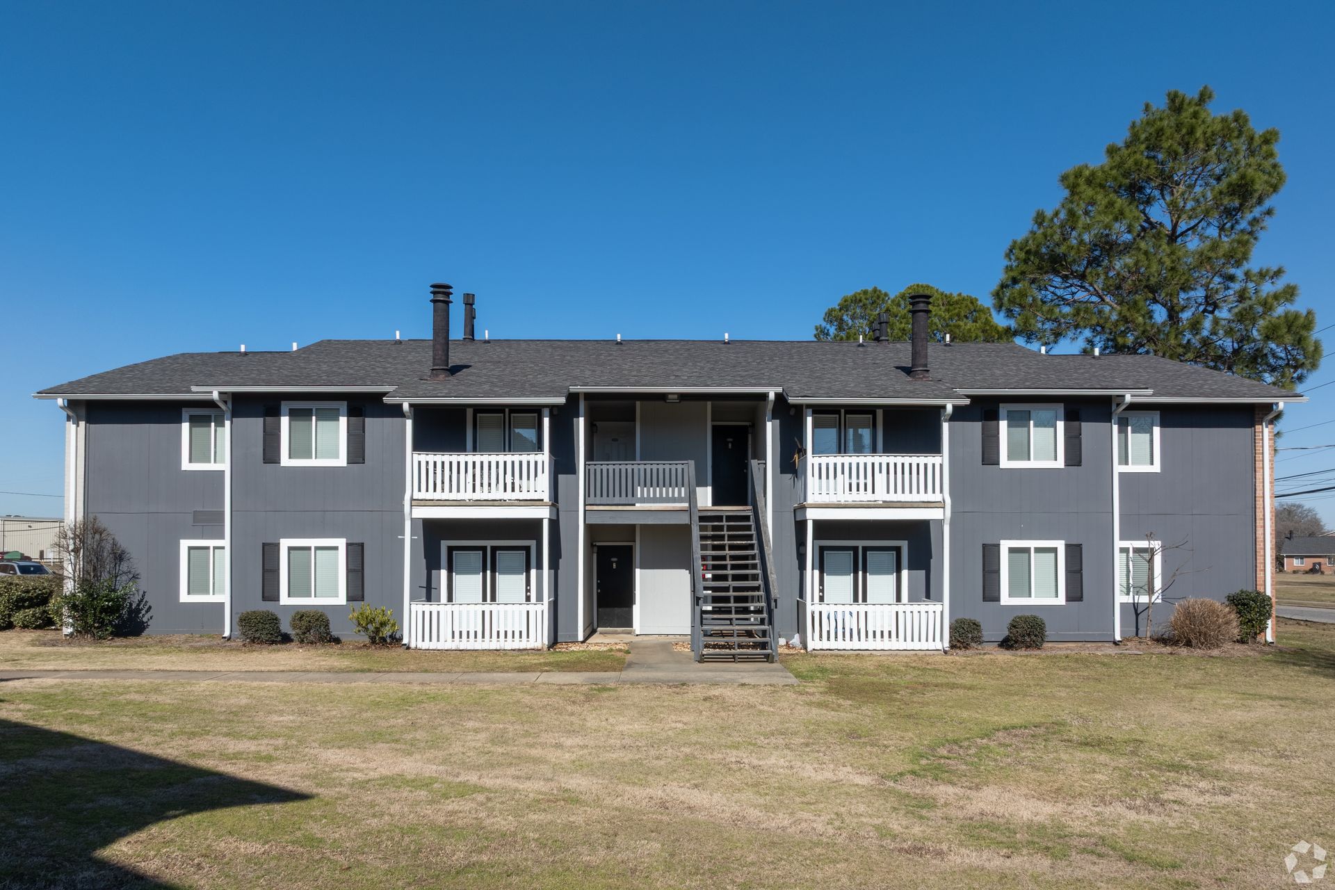 A two-story, grey apartment building with white balconies and stairs, set against a clear blue sky.