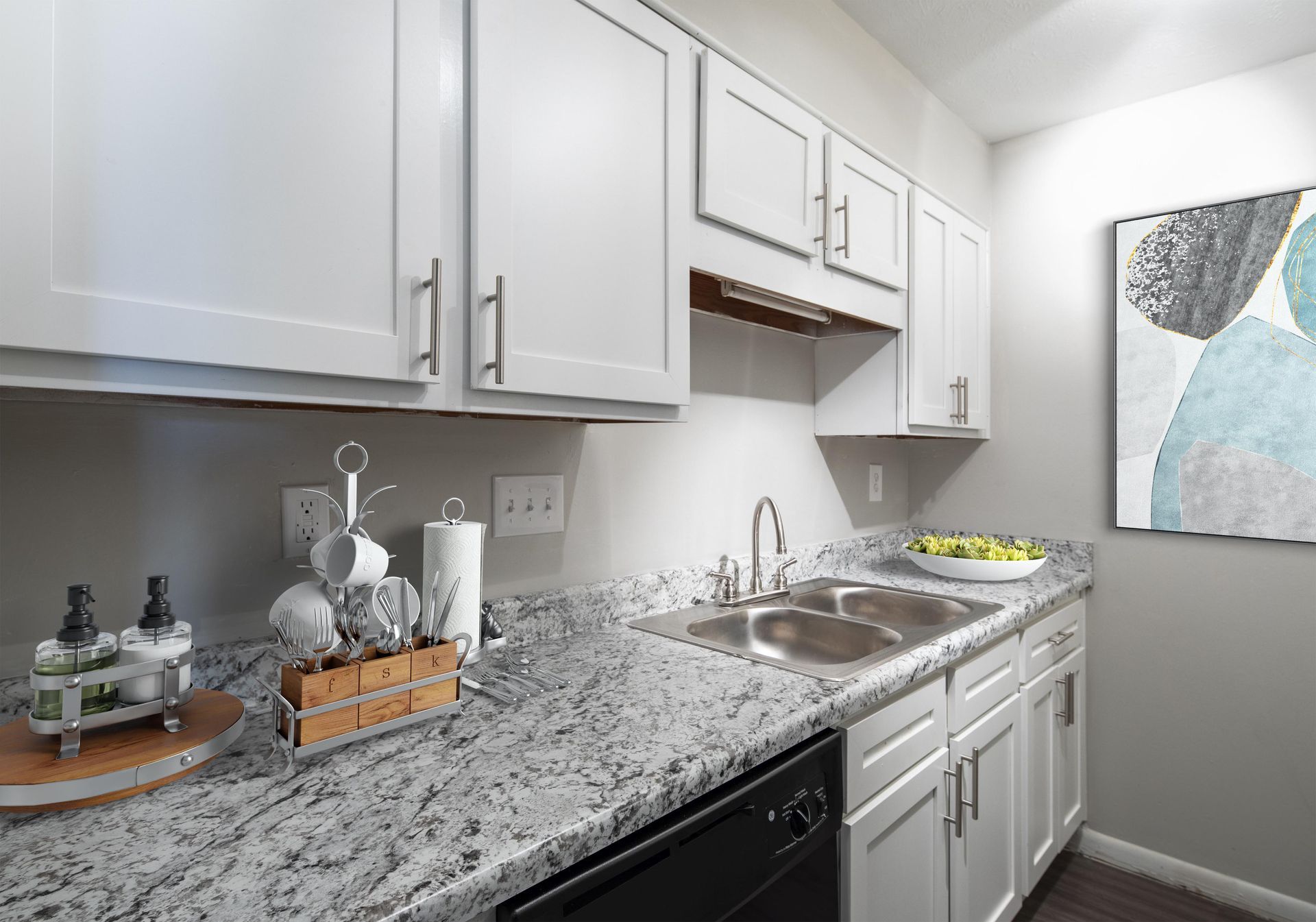 A kitchen with white cabinets, granite countertops, a double sink, black dishwasher, and abstract wall art.