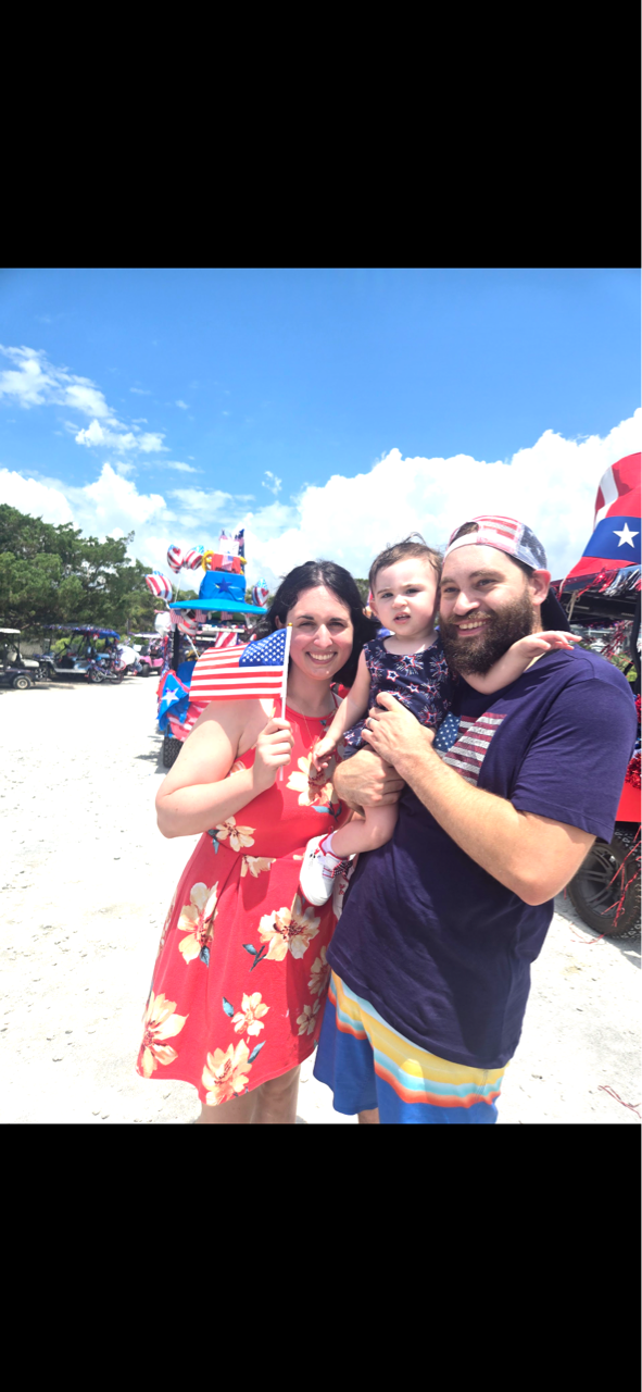 Family of three posing with American flags, smiling under a blue sky. Mother in a red floral dress, father with a beard, holding a baby.