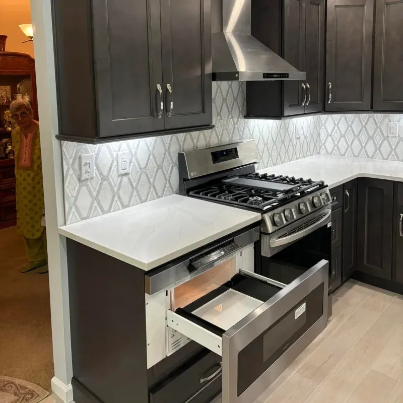 A modern kitchen with dark gray cabinets, white countertops, and a stainless steel range and microwave drawer. An older woman stands in the background.