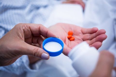 Hands holding a blue bottle cap with white liquid and orange pills over a hospital bed