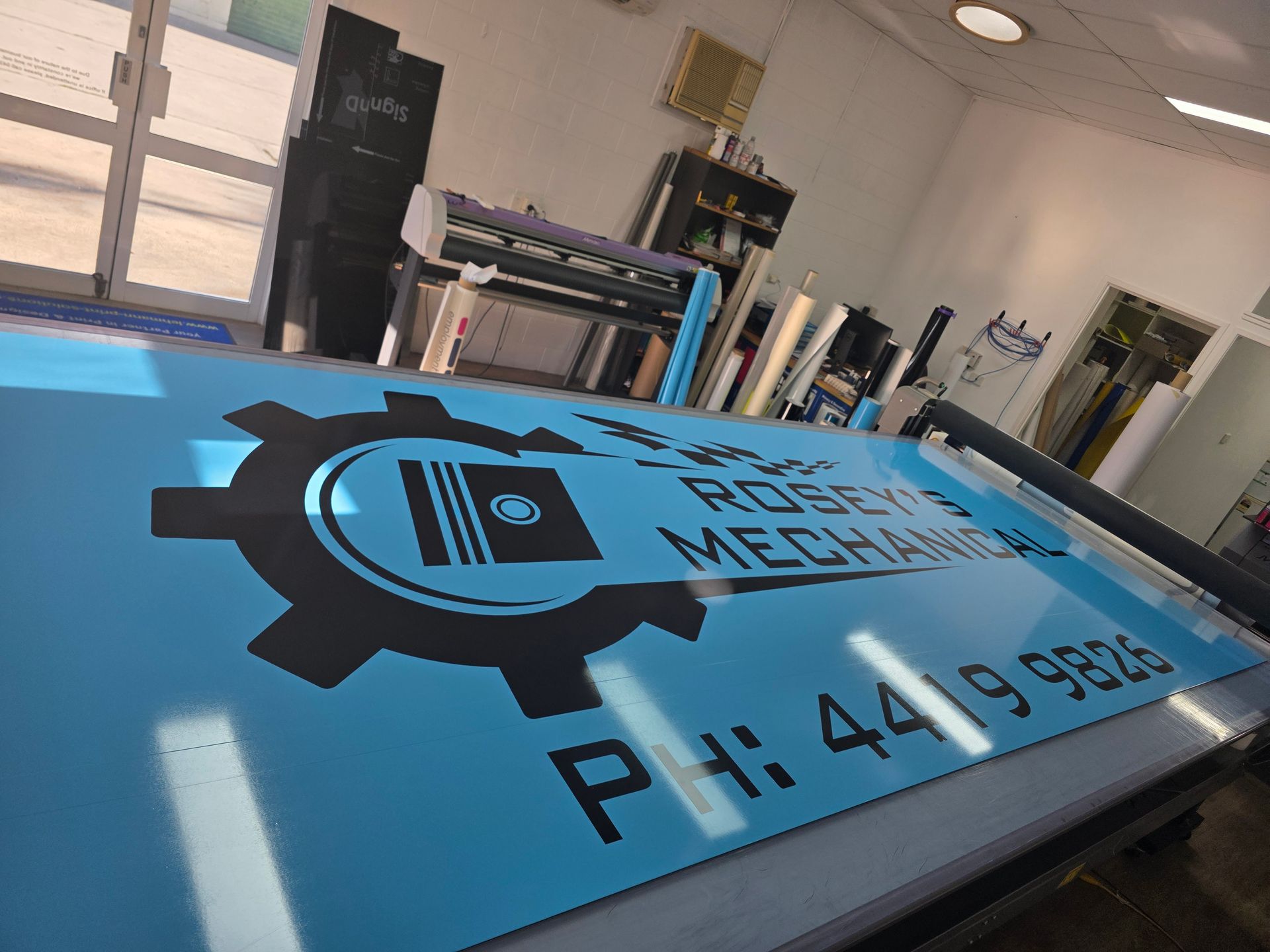 Blue Sign With Gear Logo and Phone Number Inside a Workshop — Lehmann Print Solutions in Bowen, QLD