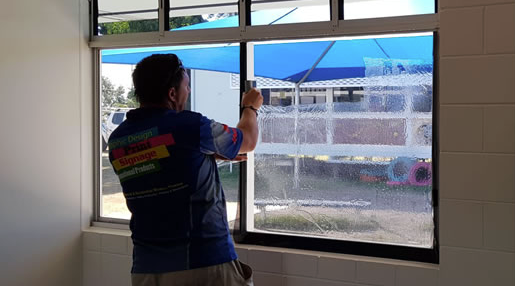 Person applying window film to a large glass window in a bright room. — Lehmann Print Solutions in Bowen, QLD