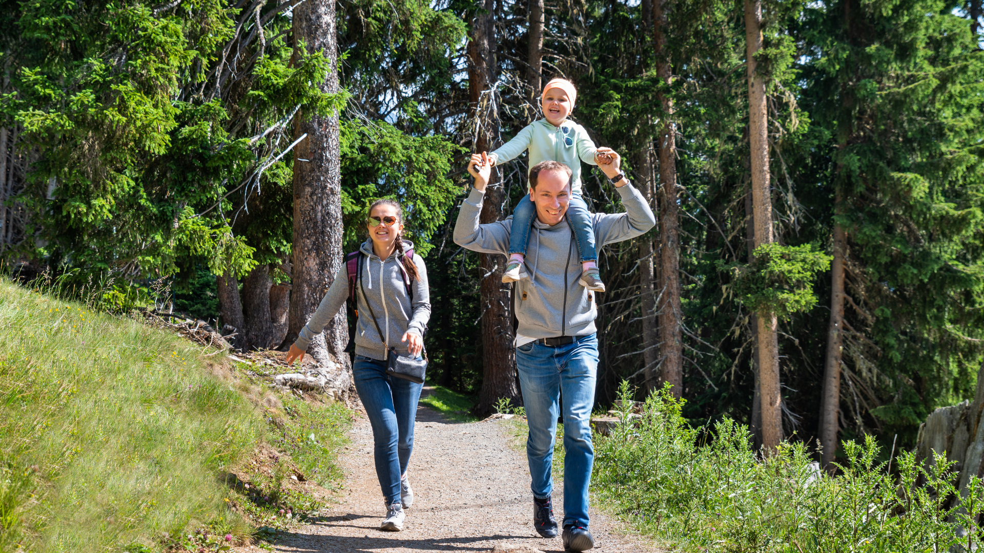 Two hikers on a forest trail, one carrying a child on his shoulders under tall trees.