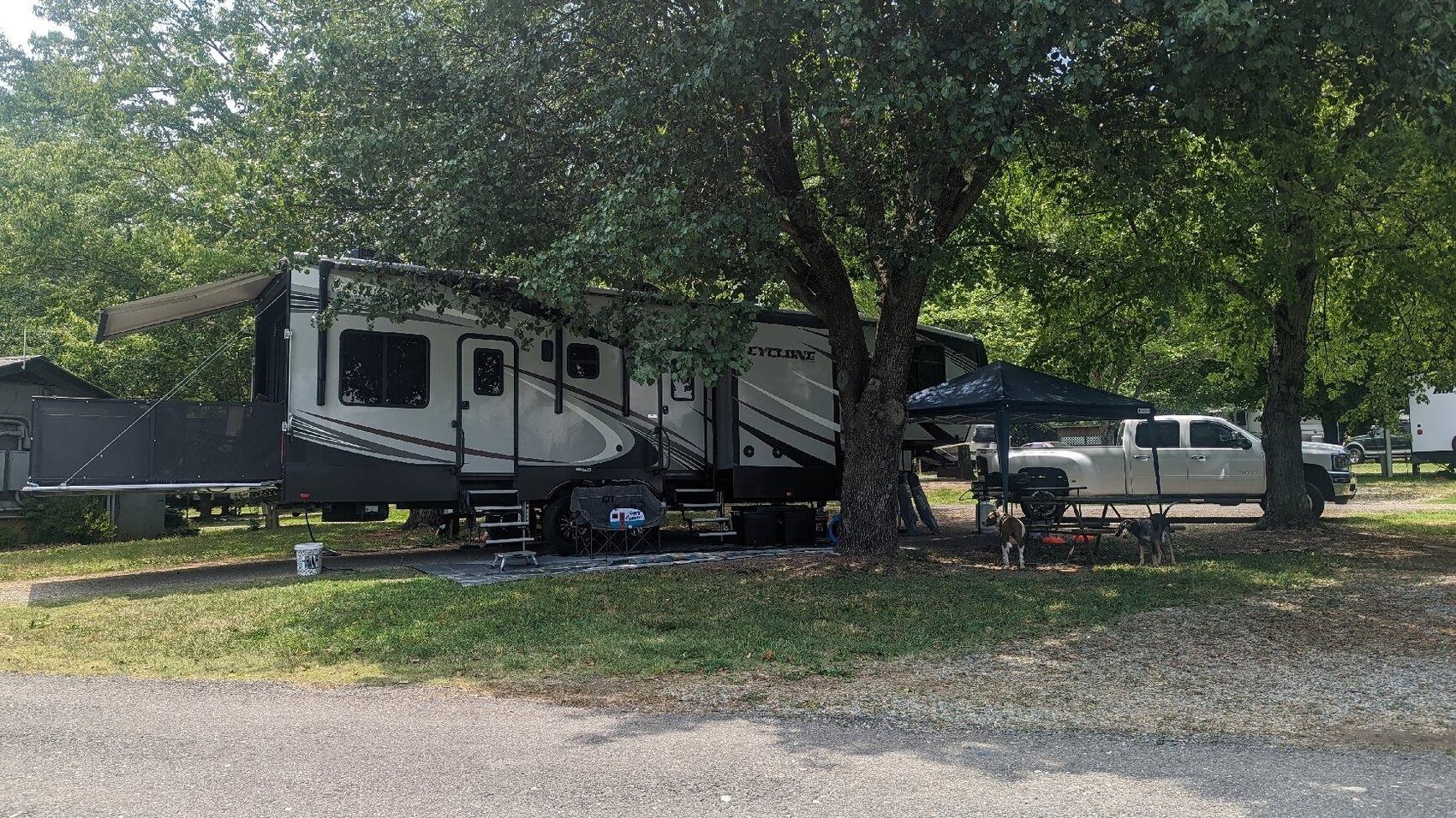 Camper trailer parked under trees beside a gravel road in a shaded campground
