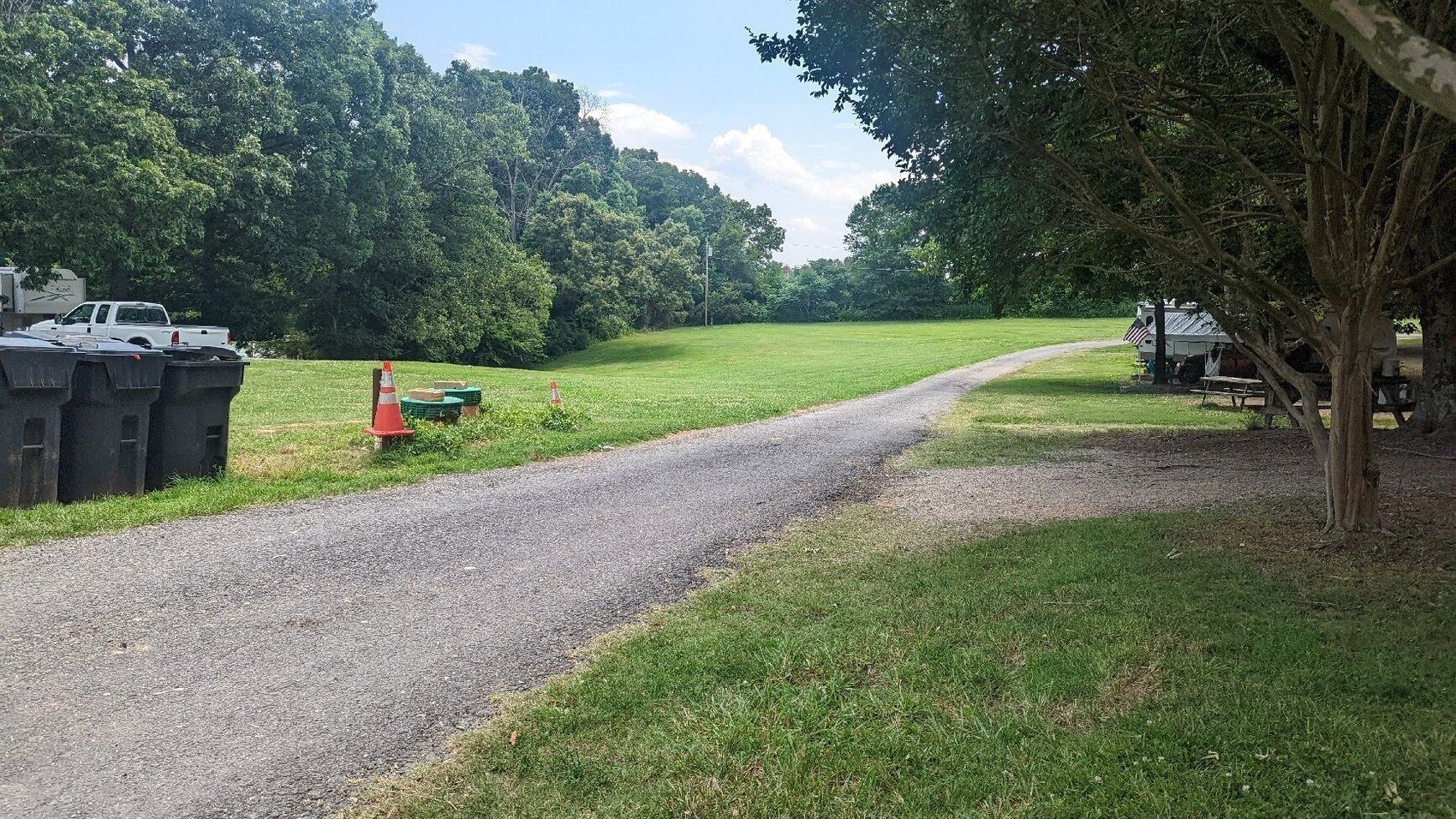 Gravel path curving through a grassy park with trees and trash bins along the left side.
