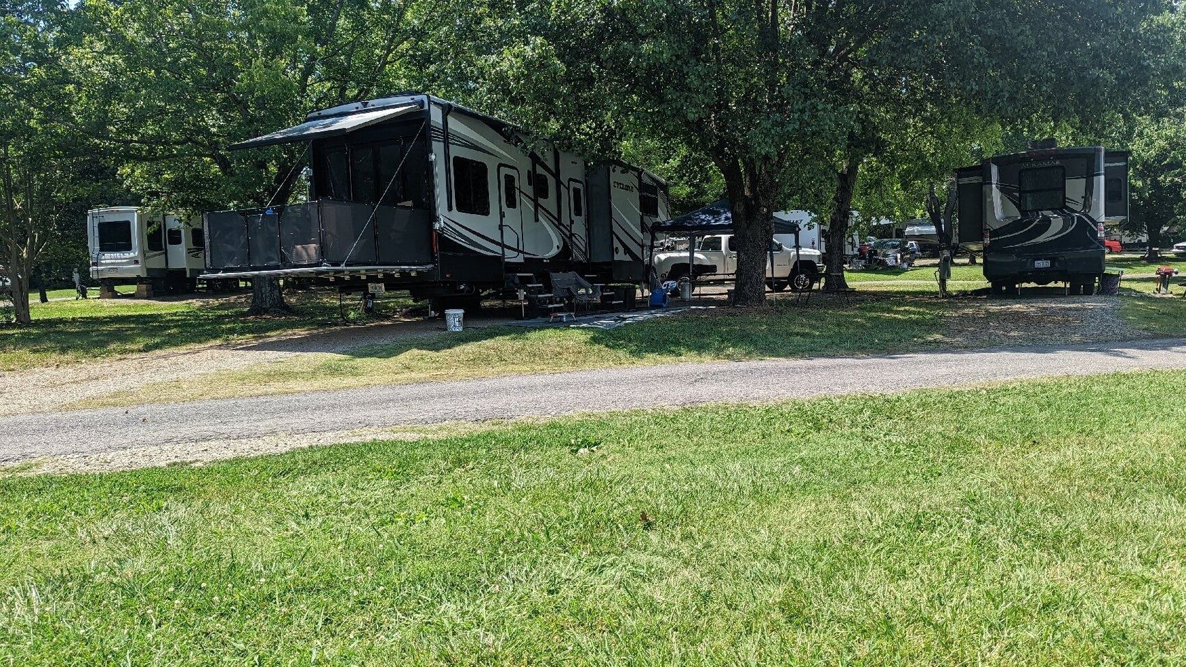 A large black RV parked on a grassy campsite under trees, with another RV in the background.