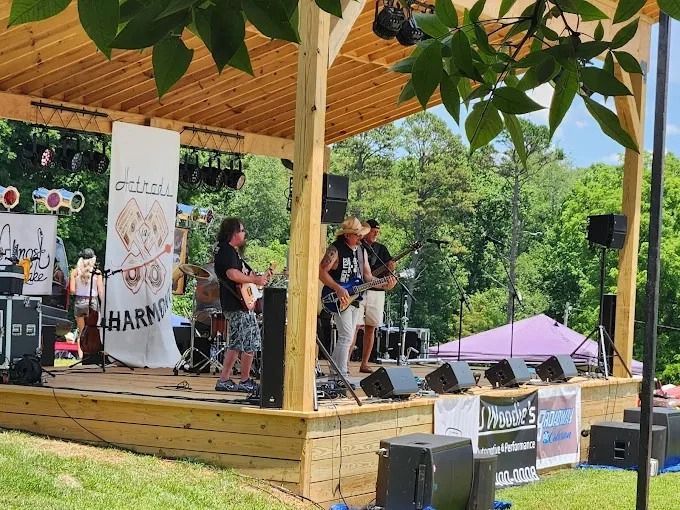 Outdoor live band performing on a wooden stage under a pavilion, with trees and speakers around them.