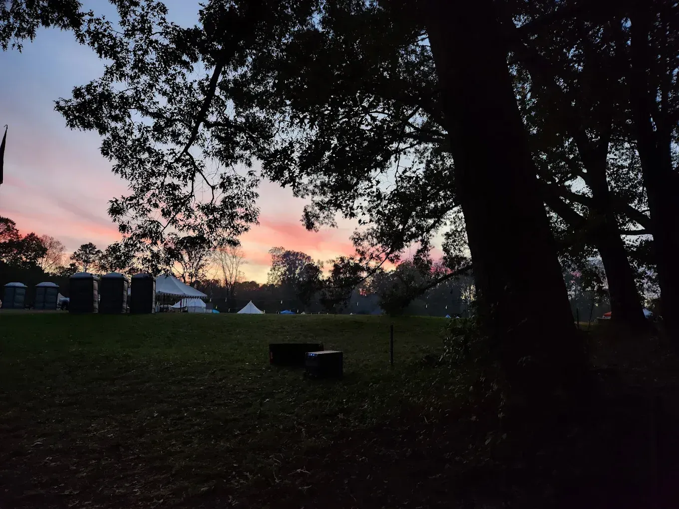 Sunset over a park with silhouetted trees, grassy field, and tents or vehicles in the distance