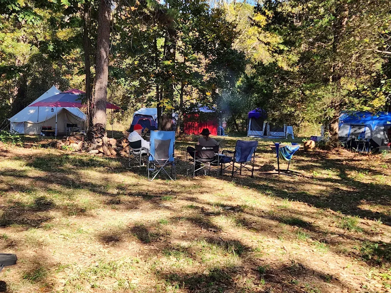 Camping area with tents and folding chairs under trees in a sunny grassy clearing