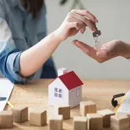 Hands exchanging keys above a small model house with toy blocks on a table