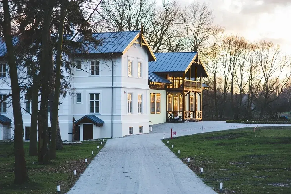 White two-story house with blue roof beside a gravel driveway and trees at dusk