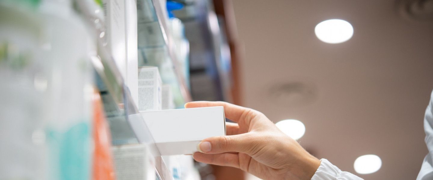 A hand reaching for a white box on a pharmacy shelf, under bright lights.