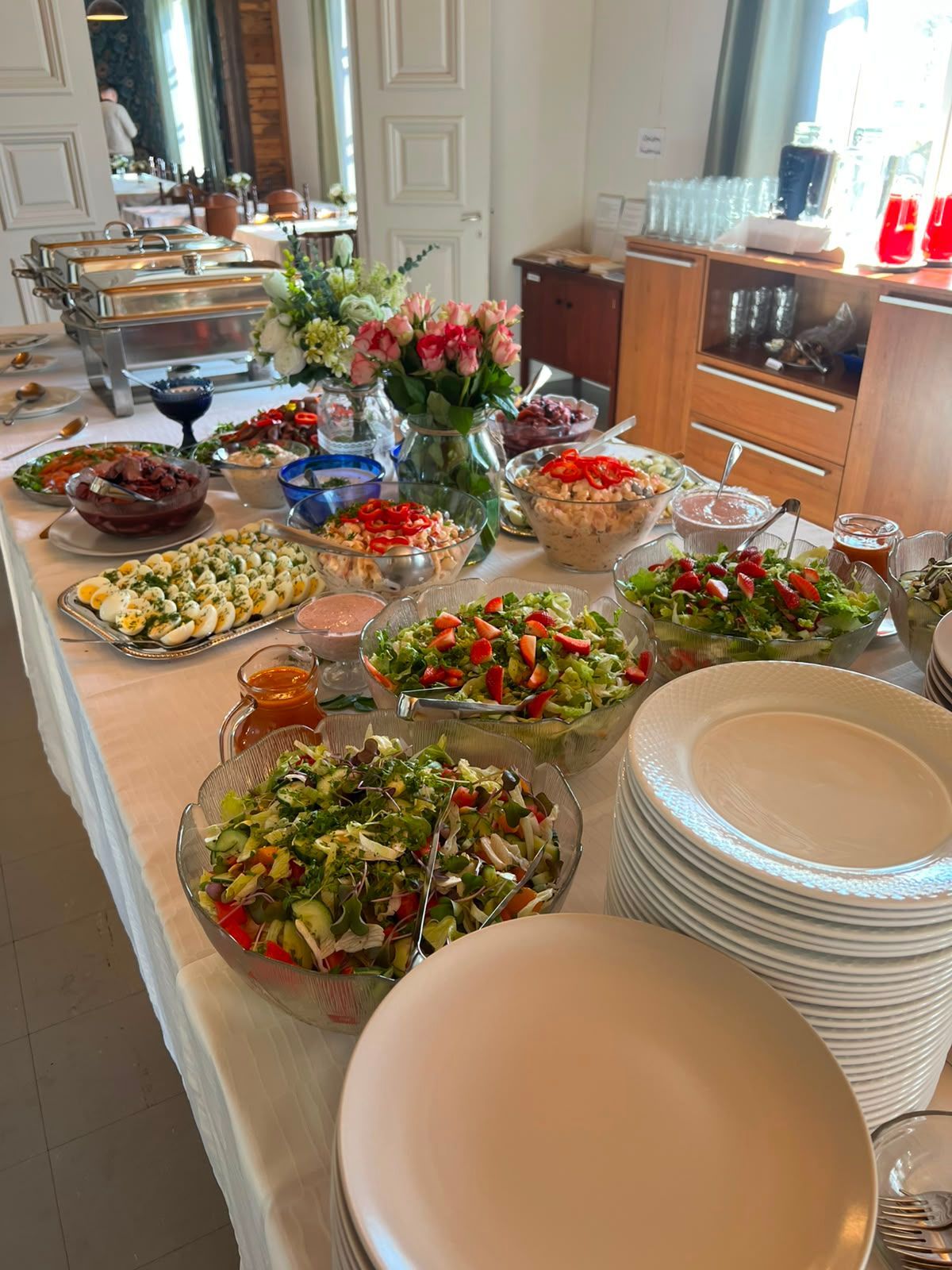 Buffetpöytä, jossa on salaatteja, leipää, hedelmiä ja pinottuja lautasia valoisassa ruokasalissa.