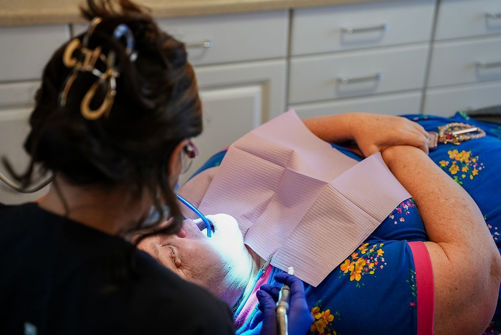 Dentist treating a reclining patient in a clinic, with a bright examination light and pink protective bib.