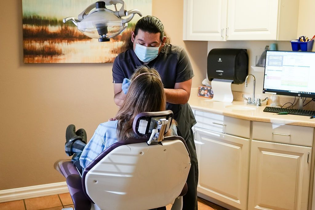 Dentist examining a patient in a dental chair, with clinic equipment and computer monitor nearby.