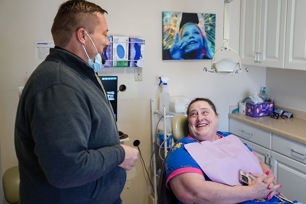 Man receiving a dental exam while seated in a clinic, smiling as the dentist stands nearby.