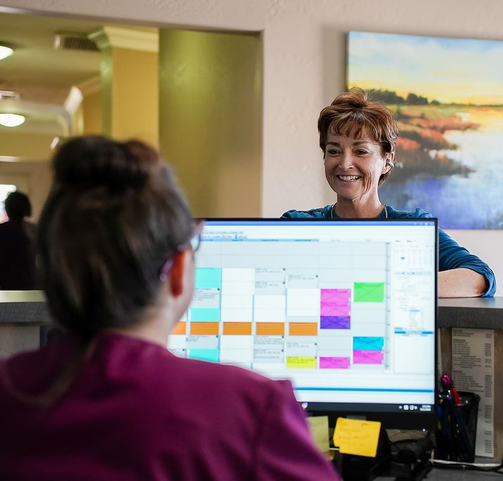 Two coworkers talking across a desk with a computer monitor showing a colorful calendar schedule
